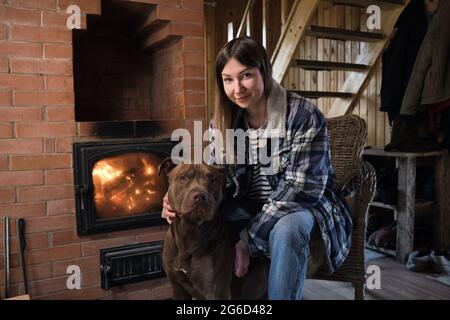 Porträt einer jungen Frau, die zusammen mit ihrem Hund in der Nähe des Kamins sitzt und die Kamera anschaut Stockfoto