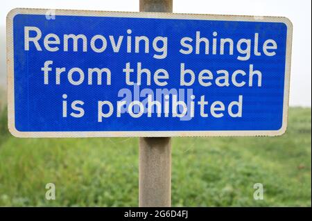 Blau-weißes Schild mit Text: Das Entfernen von Kiesel vom Strand ist verboten. Isoliert mit verschwommenem Hintergrund von Gras und neblig weißem Himmel. Stockfoto