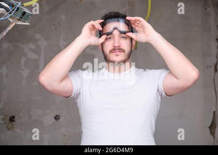 Junger Baumeister, der auf einer Baustelle eine Schutzbrille aufsetzt. Mann mit Schutzbrille. Stockfoto