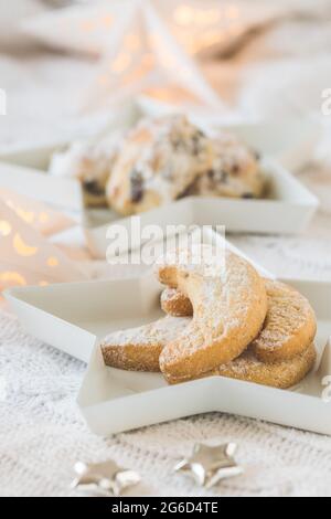 Vanillegebäck und Mini-Stollen auf weißen Sterntellern auf einer weißen Decke, verziert mit silbernen Sternen und Sternenlicht, Mini-Stollen im Hintergrund, Stockfoto