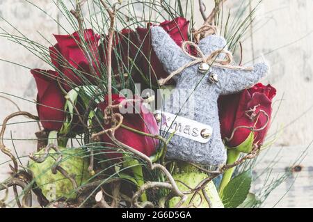 Winterlicher Rosenstrauß in einer Glasvase mit einem grauen Wollengel mit WEIHNACHTSSCHRIFT, altes Altholz als Hintergrund Stockfoto