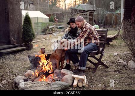 Junges Paar, das zusammen mit ihrem Hund am Feuer beim Barbecue draußen in der Nähe ihres Hauses sitzt Stockfoto