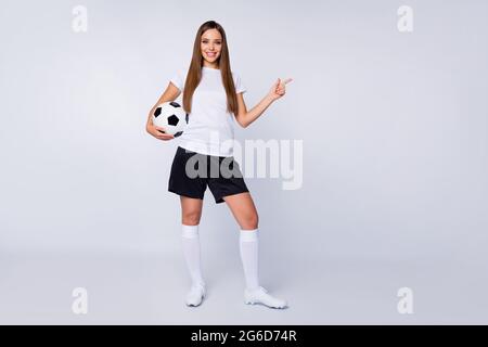 In voller Länge Foto von hübschen Kapitän Dame Spieler Fußball Team halten Lederball direkten Finger leeren Raum tragen Fußballuniform T-Shirt Shorts Stiefel Stockfoto