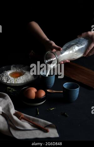 Ernteansicht der Hände einer anonymen Frau unter dramatischer Beleuchtung, die Zutaten wie Milch, Mehl und Eier zubereitet, um einen Teig zu machen Stockfoto