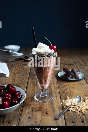 Tasse Schokoladenmilchshake mit Creme und einer Kirsche auf Holzfläche vor dunklem Hintergrund Stockfoto