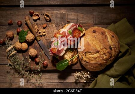 Draufsicht auf appetitlich hausgemachtes Sandwich mit frisch gebackenem hausgemachtem Brot mit geschnittenem Prosciutto und Tomaten auf rustikalem Holztisch mit Nüssen und Stockfoto
