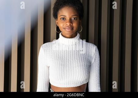 Stilvolle junge schwarze Frau im Crop-Pullover mit Blick auf die Kamera, während sie in der Nähe einer gestreiften Gebäudewand in der Stadt steht Stockfoto