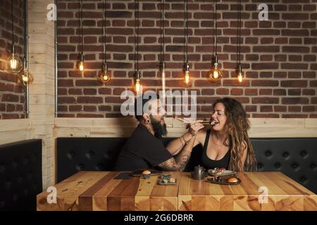 Liebende Paare essen appetitliches Sushi, während sie an einem Holztisch im japanischen Restaurant sitzen Stockfoto