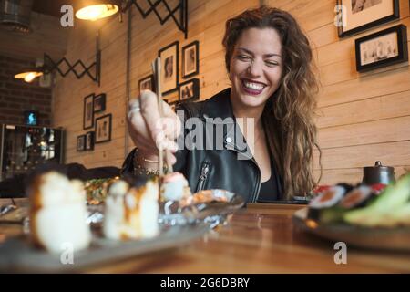 Lächelnde Frau, die im japanischen Restaurant leckeres Sushi isst, während sie am Holztisch sitzt Stockfoto