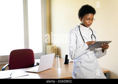 Serious junge schwarze Ärztin in einem medizinischen Mantel mit Stethoskop, die mit einer Tablette in einem modernen Klinikbüro arbeitet Stockfoto