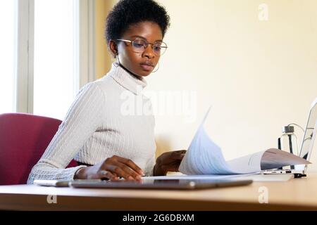 Junge afroamerikanische Ärztin, die mit einem Laptop am Tisch sitzt und Krankenakten liest, während sie im Klinikbüro arbeitet Stockfoto