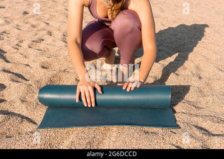 Von oben beschnitt eine nicht erkennbare junge Frau in Sportkleidung, die eine Yogamatte auf Sand legte, während sie sich auf das Training am Strand in der Nähe des Ozeans vorbereitete Stockfoto