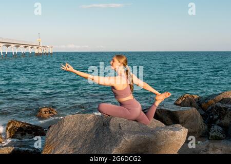 Seitenansicht einer friedlichen, flexiblen Frau, die Yoga in Eka Pada Rajakapotasana auf Felsen am Strand praktiziert Stockfoto