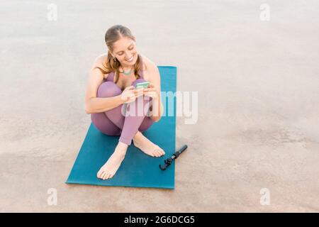 Entzückte Frau im aktiven Ohr, die auf einer Yogamatte sitzt und am Strand auf dem Mobiltelefon surft Stockfoto