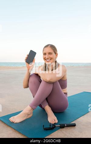 Entzückte Frau im aktiven Ohr, die auf einer Yogamatte sitzt und das Mobiltelefon am Strand vorführt Stockfoto