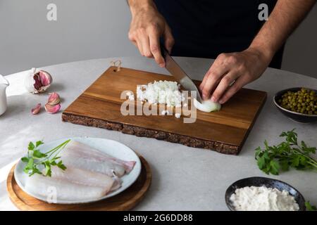 Unkenntlich kocht der Mann beim Schneiden von Zwiebeln mit einem Messer auf einem Holzbrett in der Nähe von Erbsen und Seehechtfilet mit Kräutern während der Zubereitung der Speisen in der Küche Stockfoto