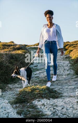 Ganzer Körper einer glücklichen ethnischen Frau mit Border Collie Hund, der am sonnigen Frühlingsabend auf dem Weg zwischen grasbewachsenen Hügeln zusammenläuft Stockfoto