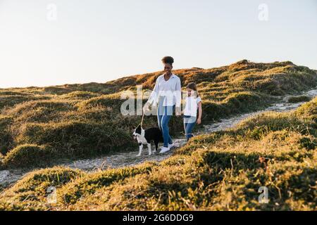 Ganzer Körper von glücklicher multiethnischer Frau und kleinem Mädchen mit Border Collie Hund, der am sonnigen Frühlingsabend gemeinsam auf dem Trail zwischen grasbewachsenen Hügeln läuft Stockfoto