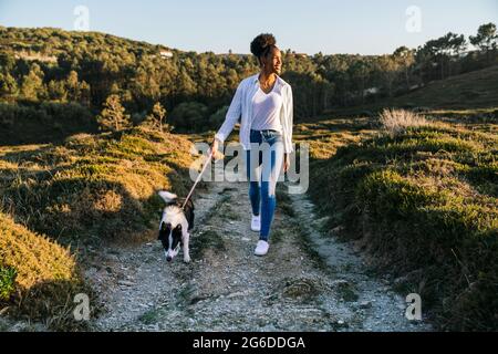 Ganzer Körper einer glücklichen ethnischen Frau mit Border Collie Hund, der am sonnigen Frühlingsabend auf dem Weg zwischen grasbewachsenen Hügeln zusammenläuft Stockfoto