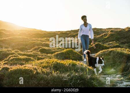 Ganzer Körper einer glücklichen ethnischen Frau mit Border Collie Hund, der am sonnigen Frühlingsabend auf dem Weg zwischen grasbewachsenen Hügeln zusammenläuft Stockfoto