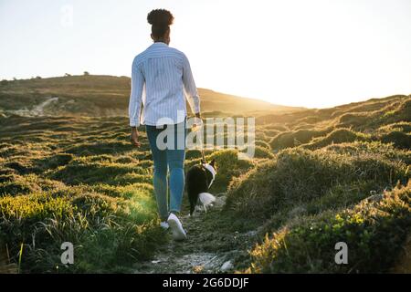 Ganzkörper-Rückansicht einer ethnischen Frau mit Border Collie Hund, der am sonnigen Frühlingsabend auf dem Weg zwischen grasbewachsenen Hügeln zusammenläuft Stockfoto