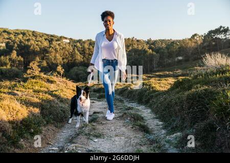 Ganzer Körper einer glücklichen ethnischen Frau mit Border Collie Hund, der am sonnigen Frühlingsabend auf dem Weg zwischen grasbewachsenen Hügeln zusammenläuft Stockfoto