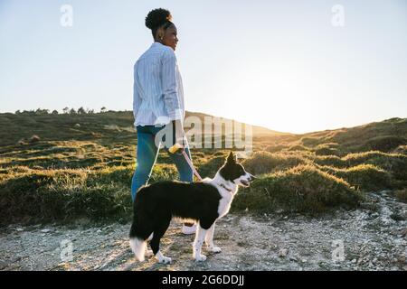 Ganzkörperrückansicht einer glücklichen ethnischen Frau mit dem Border Collie-Hund, der am sonnigen Frühlingsabend auf dem Weg zwischen grasbewachsenen Hügeln zusammenläuft Stockfoto