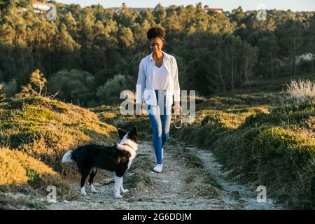 Ganzer Körper einer glücklichen ethnischen Frau mit Border Collie Hund, der am sonnigen Frühlingsabend auf dem Weg zwischen grasbewachsenen Hügeln zusammenläuft Stockfoto