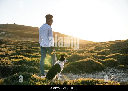 Ganzkörperrückansicht einer glücklichen ethnischen Frau mit dem Border Collie-Hund, der am sonnigen Frühlingsabend auf dem Weg zwischen grasbewachsenen Hügeln zusammenläuft Stockfoto