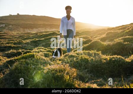 Ganzer Körper einer glücklichen ethnischen Frau mit Border Collie Hund, der am sonnigen Frühlingsabend auf dem Weg zwischen grasbewachsenen Hügeln zusammenläuft Stockfoto