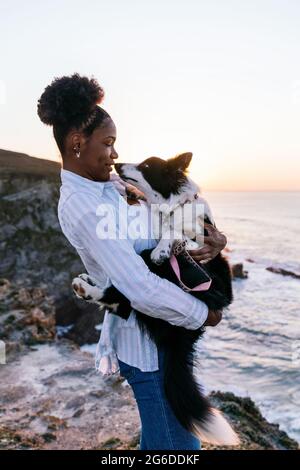 Seitenansicht der afroamerikanischen Besitzerin mit niedlichem Happy Border Collie Hund, während sie bei Sonnenuntergang in der Nähe des winkenden Meeres Zeit zusammen genießt Stockfoto