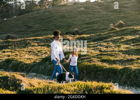 Ganzkörper-Seitenansicht einer glücklichen multiethnischen Frau und eines kleinen Mädchens mit Border Collie-Hund, der am sonnigen Frühlingsabend auf dem Weg zwischen grasbewachsenen Hügeln zusammenläuft Stockfoto