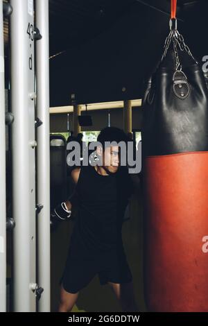 Junge fokussierte asiatische Mann Training Boxen Durchführung Schläge beim Training mit schweren Boxsack in einem modernen Fitnessstudio Stockfoto