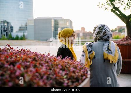 Freute Muslin-Freundinnen in Hijabs, die auf der Bank saßen und einander anschauten, während sie ein Wochenende in der Stadt verbrachten Stockfoto