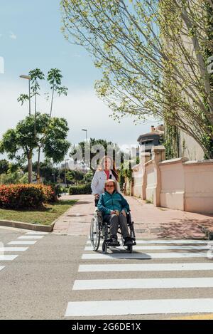Erwachsene Frau, die im Sommer bei einem Spaziergang den Rollstuhl mit der älteren Mutter schiebt und die Straße in der Stadt überquert Stockfoto