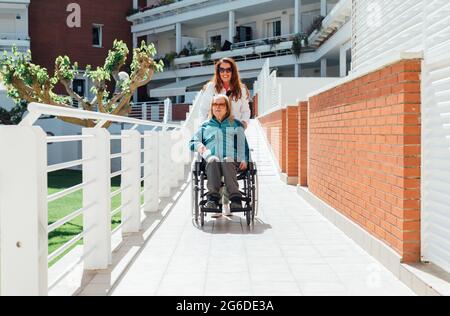 Glückliche Erwachsene Tochter schieben Rollstuhl mit älteren Mutter, während Spaziergang am sonnigen Tag genießen Stockfoto