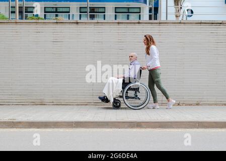 Seitenansicht einer erwachsenen Tochter, die mit einem älteren Vater den Rollstuhl schiebt, während sie zusammen in der Stadt läuft Stockfoto