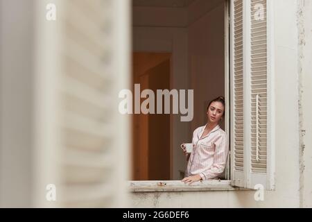 Ruhige Frau im Pyjama, die bei einer Tasse Kaffee am Morgen in der Nähe des Fensters steht und wegschaut Stockfoto