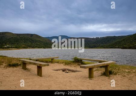 Vier Holzbänke an einem Strand mit Wasser und Hügeln im Hintergrund. Stockfoto