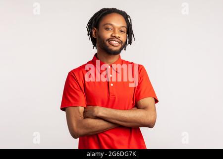 Junger erwachsener bärtiger Mann mit Dreadlocks in einem roten, lässigen T-Shirt, der mit selbstbewusster Gesichtsausdruck und gekreuzten Armen posiert und lächelnd auf die Kamera blickt Stockfoto