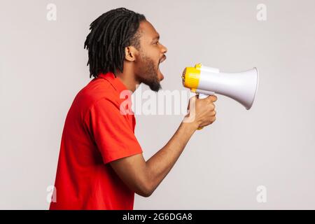 Das Profil eines Mannes mit Dreadlocks und Bart, der ein rotes T-Shirt im lässigen Stil trägt, laut nach Megaphon schreit, protestiert, will gehört werden. Indoor st Stockfoto