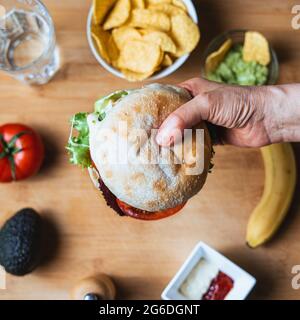 Frischer, leckerer Burger mit rustikalem Brötchen, umgeben von Nachos mit Guacamole zum Eintauchen, ein Wasserglas, eine Tomate, eine Avocado, Pfeffer, Ketchup und Mayonnaise Stockfoto