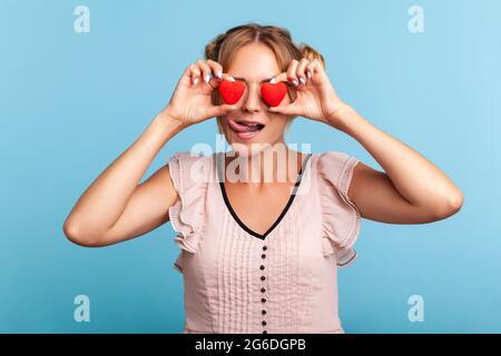 Liebende Augen. Schöne junge Erwachsene Frau mit zwei lustigen Haarbrötchen, die wie eine Brille zwei valentinsherzen vor ihren Augen halten, zeigt die Zunge heraus. Zoll Stockfoto