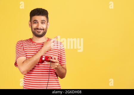Lächelnder positiver männlicher Gamer mit Bart in gestreiftem T-Shirt, der Gamepad hält und mit dem Finger auf leeren Raum zeigt, Werbung für Videospiele-Wettbewerb. Indo Stockfoto