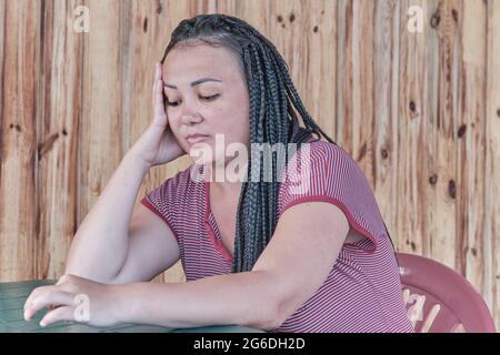 Porträt einer jungen, schönen asiatischen Frau, mit Dreadlocks auf ihren schwarzen Haaren, in legerer Kleidung, an einem Tisch sitzend und mit ha Stockfoto