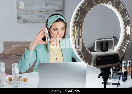 Stilvolle Frau zeigt Sprühflasche mit mizellarem Wasser in der Nähe von Laptop und Telefonhalterung mit Ringlampe Stockfoto