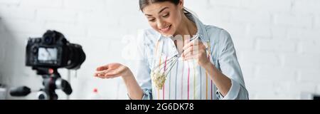 Fröhliche kulinarische Blogger hält Schneebesen mit rohem Teig in der Nähe verschwommene Digitalkamera, Banner Stockfoto