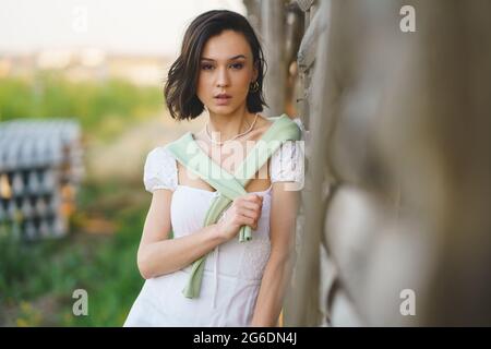 Asiatische Frau, die in der Nähe eines Tabaktrocknungsschuppens posiert und ein weißes Kleid und grüne Gummistiefel trägt. Stockfoto