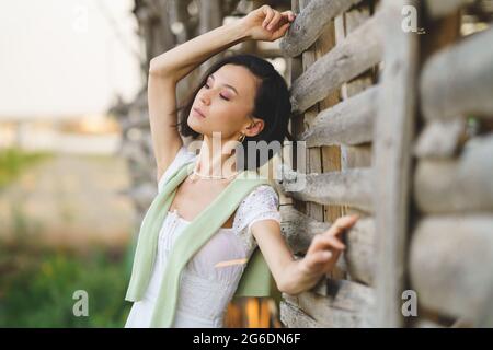 Asiatische Frau, die in der Nähe eines Tabaktrocknungsschuppens posiert und ein weißes Kleid und grüne Gummistiefel trägt. Stockfoto