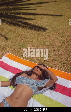 Vertikale Ansicht schwarze indische Womanliegend in einem Handtuch auf dem Gras mit Palmen Schatten auf ihrem Körper Sonnenbaden nach einem Bad im Pool. Summer Vibes 2021. Stockfoto